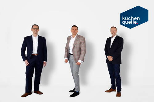 Drei Geschäftsmänner in Anzügen posieren vor weißem Hintergrund neben dem blauen Logo von küchenquelle
