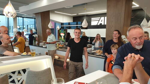 Fröhliche Gruppe von Kolleginnen, Kollegen und Kindern bei einem lockeren Buffet-Empfang in einer modernen Büro-Küche, während ein junger Mann im schwarzen T‑Shirt lächelnd im Zentrum steht