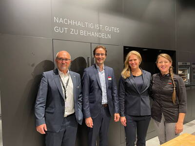 Vier lächelnde Personen in Business- und Kochkleidung posieren vor einer dunklen Küchenwand mit dem Schriftzug „Nachhaltig ist, Gutes gut zu behandeln“ in einer modernen Ausstellungsküche.