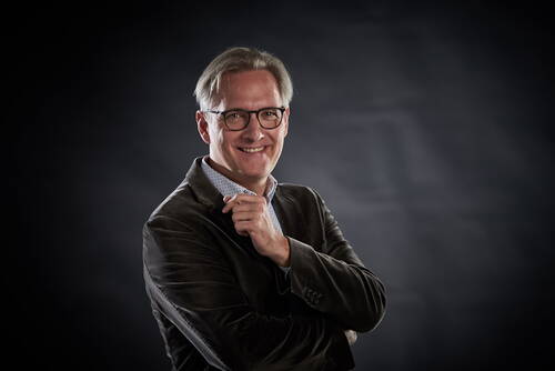 Lächelnder Mann mittleren Alters mit Brille und grauem Haar posiert im Sakko vor dunklem Studiohintergrund