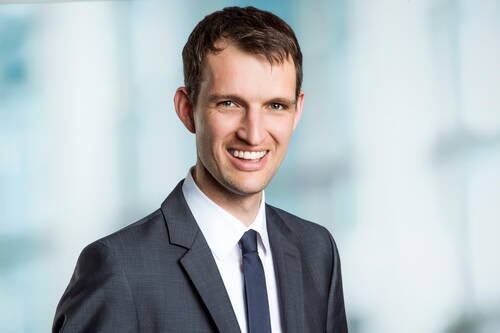 Junger Geschäftsmann im dunkelgrauen Anzug und Krawatte lächelt freundlich vor unscharfem, hellem Bürohintergrund