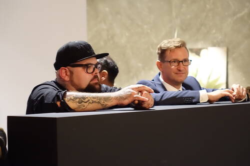 Zwei Männer lehnen entspannt an einer dunklen Theke, einer mit Basecap, Bart und tätowiertem Arm, der andere im Anzug und mit Brille, während sie aufmerksam in den Raum blicken.