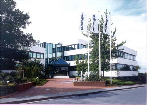 Modernes Bürogebäude mit großflächigen Glasfronten, bepflanztem Eingangsbereich und drei weißen Flaggenmasten an der Straße