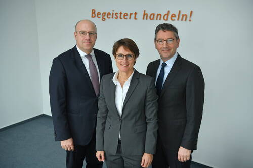 Drei geschäftlich gekleidete Personen stehen lächelnd in einem Büro vor einer weißen Wand mit dem orangefarbenen Schriftzug „Begeistert handeln!“.