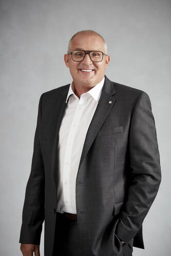 Älterer, lächelnder Mann mit Brille im dunklen Anzug und weißem Hemd steht vor neutral grauem Hintergrund und blickt direkt in die Kamera.
