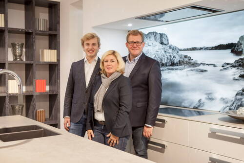 Drei elegant gekleidete Personen stehen lächelnd in einer modernen Küche vor einer Wand mit großformatigem Küstenlandschaftsbild