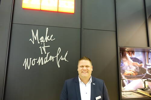 Lächelnder Messemitarbeiter im Anzug steht vor dunkler Wand mit dem Schriftzug „Make it wonderful“ und beleuchtetem Küchenmotiv im Hintergrund