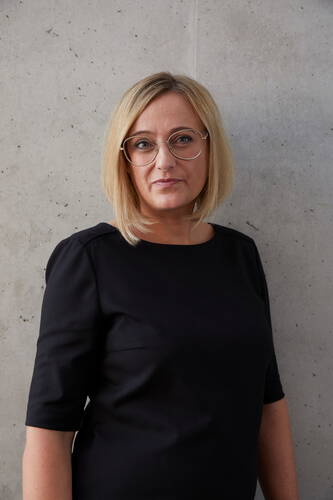 Frau mit schulterlangem blondem Haar und runder Brille steht in schwarzem Kleid vor einer grauen Betonwand und blickt ruhig in die Kamera