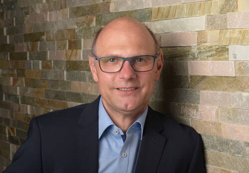 Mittelalterlicher Mann mit Brille im blauen Anzug lächelt vor einer Wand aus Natursteinplatten