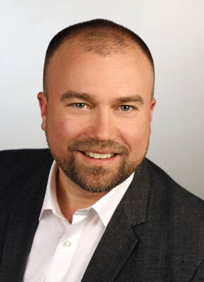 Lächelnder, bärtiger Mann im Anzug mit weißem Hemd vor neutral hellem Hintergrund in einem professionellen Portrait.
