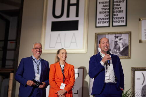 Drei lächelnde Konferenzteilnehmende im Business-Outfit stehen auf einer Bühne, während der rechte Mann mit Mikrofon ins Publikum spricht und hinter ihnen gerahmte Typografie-Plakate hängen.