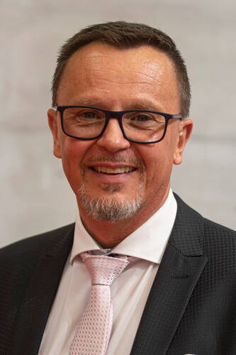Lächelnder Mann mittleren Alters mit Brille, kurzem braunen Haar und grauem Kinnbart in dunklem Anzug, weißem Hemd und rosa Krawatte vor neutralem Hintergrund