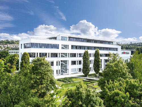 Modernes, weißes Bürogebäude mit großflächigen Glasfassaden, umgeben von dichtem Grün und Bäumen unter blauem Himmel mit einzelnen Wolken