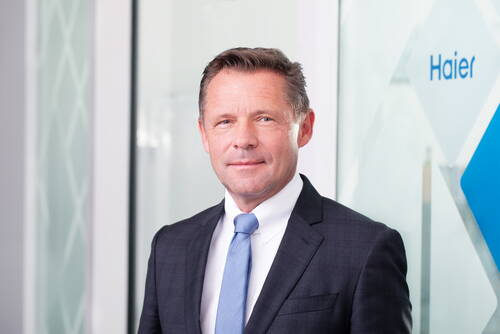Geschäftsmann mittleren Alters im dunklen Anzug mit hellblauer Krawatte steht lächelnd in einem modernen Büro vor einer Glaswand mit Haier-Logo