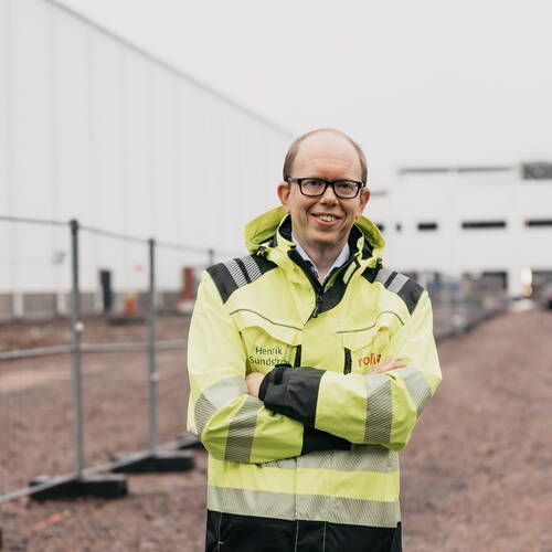 Lächelnder Bauleiter in neongelber Warnjacke steht mit verschränkten Armen vor einer großen Industriehalle auf einer Baustelle.