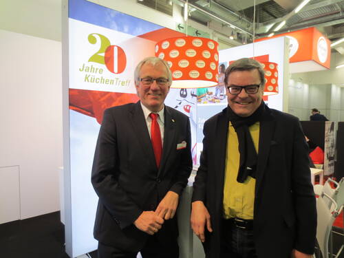 Zwei lächelnde Männer im Anzug und mit Schal stehen auf einer Messe vor einem KüchenTreff‑Stand mit dem Schriftzug „20 Jahre KüchenTreff“.