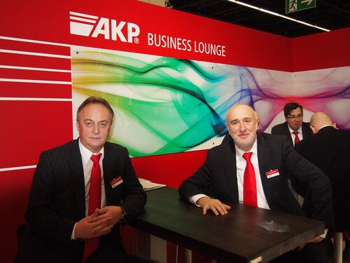 Zwei geschäftsmänner im Anzug mit roten Krawatten sitzen an einem Stehtisch in einer rot gestalteten AKP Business Lounge auf einer Messe.