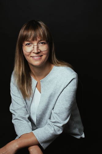 Lächelnde Frau mit langen glatten Haaren und runder Brille sitzt vor dunklem Hintergrund und trägt eine hellgraue Jacke über einer weißen Bluse
