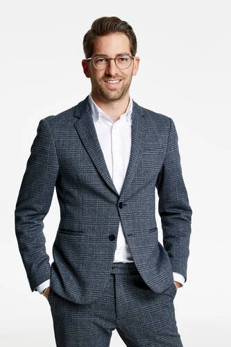Junger Mann mit Brille im grauen karierten Anzug, der mit den Händen in den Hosentaschen freundlich in die Kamera lächelt.