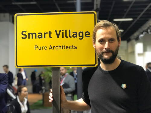 Mann mit Bart steht auf einer Messe neben einem gelben Schild mit der Aufschrift „Smart Village – Pure Architects“ und hält den Pfosten fest.