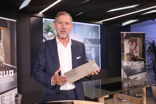 Älterer Mann im Anzug präsentiert in einer modernen Studio-Umgebung ein rechteckiges Muster einer hellgrauen Küchenarbeitsplatte und erklärt dessen Eigenschaften vor Werbebannern der Marke „alpinova“.