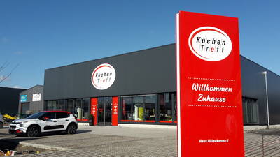 Modernes Küchenstudio „KüchenTreff“ mit großem roten Willkommensschild und geparktem Firmenauto auf dem Parkplatz.