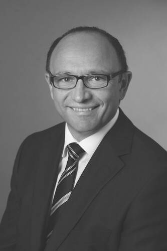 Lächelnder Mann mit Brille im Anzug und gestreifter Krawatte vor neutralem Hintergrund in schwarz-weißer Porträtaufnahme