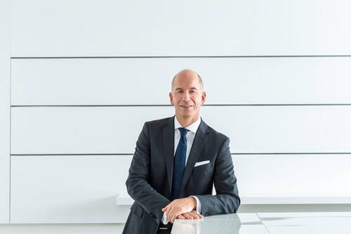 Älterer Geschäftsmann im dunklen Anzug lehnt lächelnd an einem Glastisch in einem modernen, hellen Büro mit minimalistischer Wandgestaltung