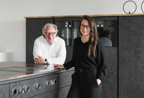 Zwei lächelnde Personen mit Brille lehnen in einer modernen, dunkel gehaltenen Küche an einer Kochinsel mit integrierten Bedienknöpfen und Einbauöfen im Hintergrund