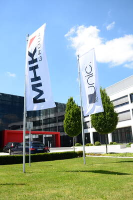 Zwei Fahnen mit den Schriftzügen „MHK Group“ und „unic.“ wehen vor einem modernen Bürogebäude mit Glasfassade, rotem Eingangsportal und gepflegtem Grünbereich unter blauem Himmel.
