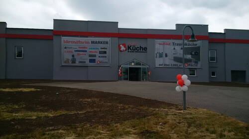 Graues Küchenfachmarkt-Gebäude mit großen Werbebannern und rot-weißen Luftballons am Eingang und an einer Straßenlaterne