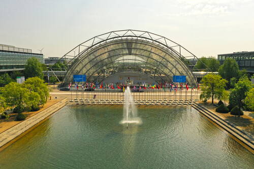 Moderne Glashallen-Eingangsarchitektur einer Messe mit Springbrunnen im Vordergrund und einer Reihe internationaler Flaggen entlang des Wasserbeckens