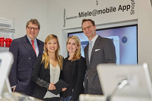 Vier Geschäftsleute stehen lächelnd vor einem Bildschirm am Miele@mobile App Messestand und posieren für ein Gruppenfoto.