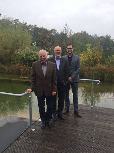 Drei Männer in Anzügen stehen lächelnd auf einem Holzsteg vor einem Teich mit herbstlich gefärbten Bäumen im Hintergrund
