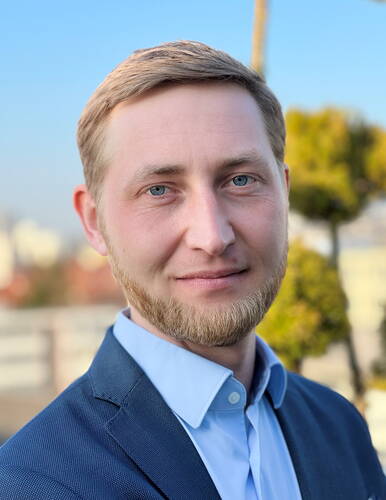 Junger Mann im blauen Anzug lächelt leicht in die Kamera, im Hintergrund unscharfe Stadtansicht bei klarem Himmel