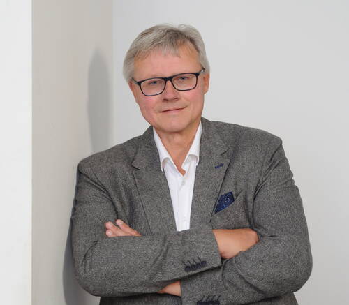 Älterer Mann mit Brille und grauem Sakko steht mit verschränkten Armen vor einer weißen Wand und blickt ruhig in die Kamera