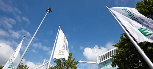 Mehrere weiße Firmenfahnen wehen vor blauem Himmel neben einem modernen Messegebäude mit Stahl- und Glaselementen.