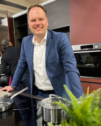 Mann im blauen Anzug lehnt lächelnd an einer modernen Kücheninsel mit Induktionskochfeld und Töpfen im Vordergrund.