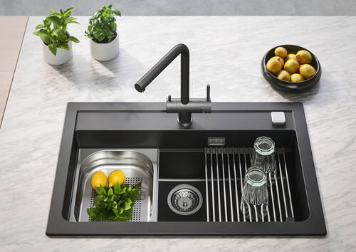 Moderne schwarze Küchenspüle auf Marmorplatte mit hohem Wasserhahn, Abtropfgitter mit zwei Gläsern, Einsatz mit Zitronen und frischer Minze sowie Kräutertöpfen und Schale mit Zitronen im Hintergrund