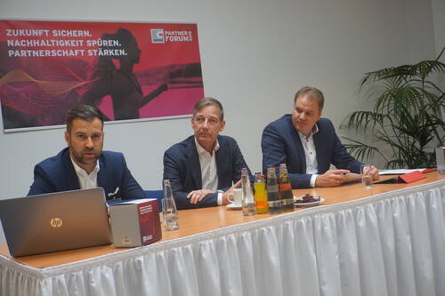 Drei Geschäftsleute im Anzug sitzen in einem Konferenzraum hinter einem mit Getränken bestückten Tisch und diskutieren vor einem Plakat mit der Aufschrift „Zukunft sichern. Nachhaltigkeit spüren. Partnerschaft stärken.“