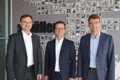 Drei lächelnde Männer im Business-Anzug stehen nebeneinander vor einer Wand mit vielen kleinen Porträtfotos von Mitarbeitenden