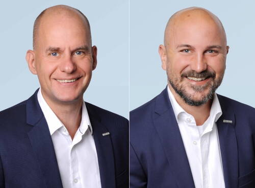 Zwei lächelnde, kahlköpfige Männer in dunklen Anzügen und weißen Hemden stehen vor hellblauem Hintergrund in einem professionellen Porträt nebeneinander.