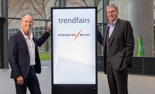 Zwei Geschäftsleute im Anzug stehen lächelnd in einer modernen Halle neben einem beleuchteten Display mit den Logos von trendfairs und NürnbergMesse.