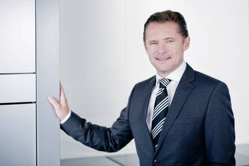 Geschäftsmann im dunklen Anzug lehnt lächelnd an einem modernen, silbernen Büroschrank in heller Umgebung