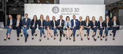 Messeteam von area30 sitzt in Businesskleidung lächelnd vor einer weißen Wand mit dem Schriftzug „area30 – design. kitchen. technics.“ nebeneinander auf einer Theke.
