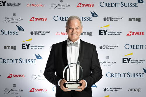 Älterer Mann im Smoking hält lächelnd eine EY-Entrepreneur-of-the-Year-Trophäe vor einer Sponsorwand mit Logos von Credit Suisse, EY und weiteren Partnern.