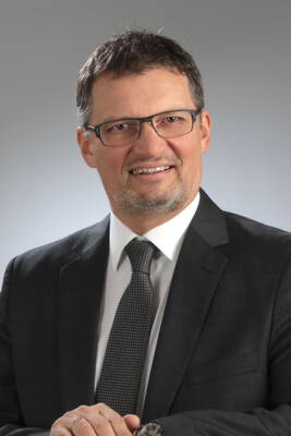 Mittelalterlicher Mann mit Brille im dunklen Anzug und gemusterter Krawatte lächelt freundlich in die Kamera vor neutral grauem Hintergrund