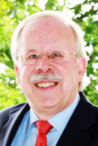Älterer Mann mit Brille, grauem Schnurrbart und roter Krawatte lächelt im Freien vor unscharfem grünen Hintergrund