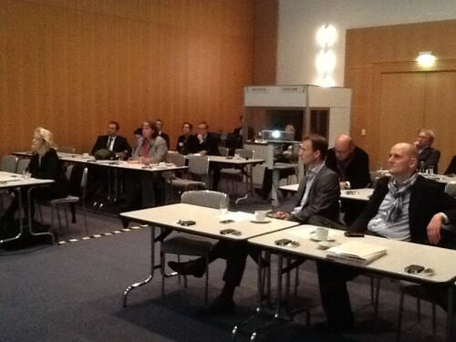 Teilnehmerinnen und Teilnehmer sitzen in einem Konferenzraum an Tischen und folgen konzentriert einer Präsentation