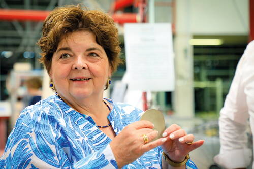 Ältere Frau in blauer gemusterter Bluse lächelt und hält eine runde Metallplatte in den Händen in einer hellen Werkshalle